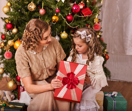 Child with mother receiving near Christmas tree. Mom gives daughter gift box with red bow.