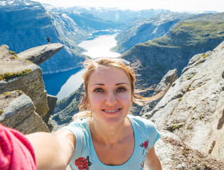 Young woman doing selfie on Trolltunga. Happy girl enjoy beautiful lake and good weather in Norway.
