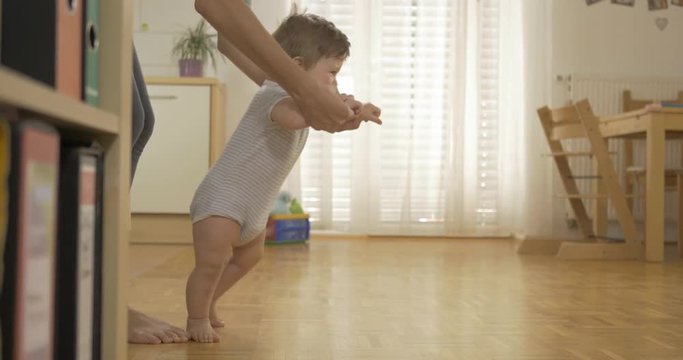 Side shot of an adorable baby boy walking barefoot across the kitchen with his mother holding him by his hands.
