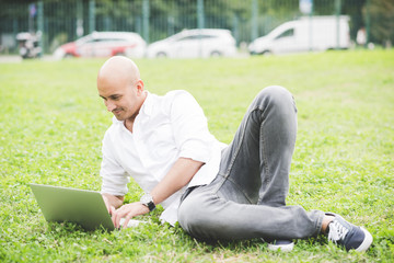 Young handsome caucasian bald business man sitting in a city park using a laptop looking down the screen laughing - working, happiness, busy concept