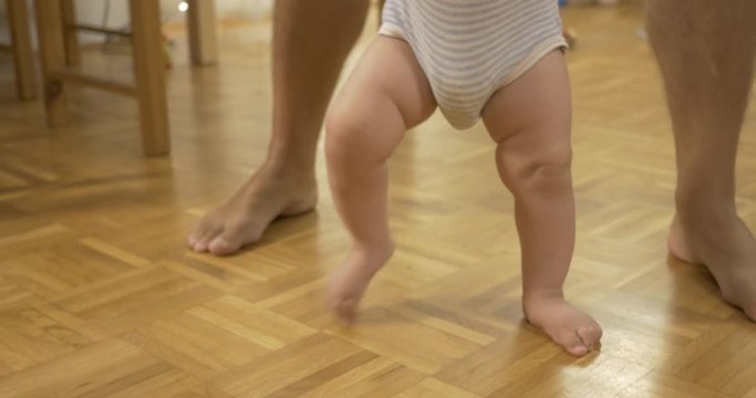 Close-up of baby's first steps at home - Baby and father walking barefoot on wooden floor.


