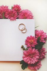 Creative Autumn background. Close-up of chrysanthemum flowers, gold wedding rings and invitation card with space for text. Autumn wedding invitation. Top view, flat layout, top view.