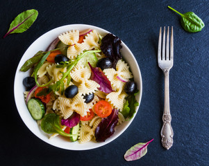 Pasta salad made from farfalle, red onion, cherry tomatoes, fresh cucumbers, black olives, mix of green salad leaves. White bowl on black stone background, top view.