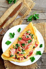 Delicious stuffed omelet on a plate. Stuffed omelet with fried sausages, grated cheese and parsley, fork, knife on an old wooden background. Breakfast recipe with eggs. Top view