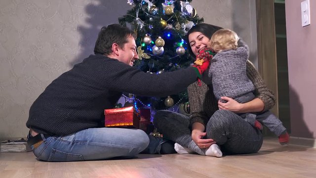 Young happy joyful family. Mother, father and baby boy son dressed in sweaters having fun near Christmas and New Year tree together at home, smile and laugh. 4k