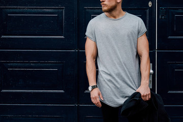 A cropped photo of a handsome muscular man wearing blank gray t-shirt while standing beside the dark blue entrance door on a street. Empty place for the logo or design. Mock up.