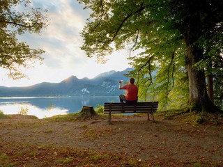 Man sit on wooden bench at mountain lake . Bank under beeches tree