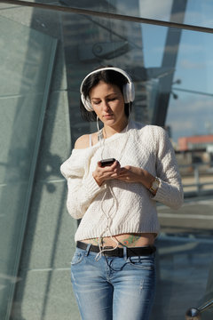 Woman in white earphones listen music standing at glassy building