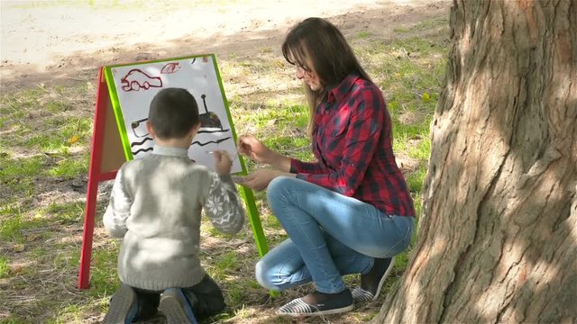 Boy and his mother draws paints on canvas and smiling. Mother and son paint on the paper. Young mother with her son drawing on the easel under a big tree