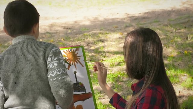 Happy mother and son having fun. Close up. Mother and son paint on the paper. Young mother with her son drawing on the easel under a big tree