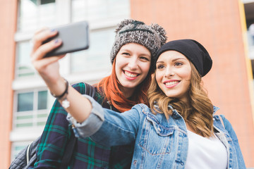 Half length of two young handsome caucasian blonde and redhead hair women hugging outdoor in the city, holding smartphone, taking selfie, smiling - social network, communication, vanity concept