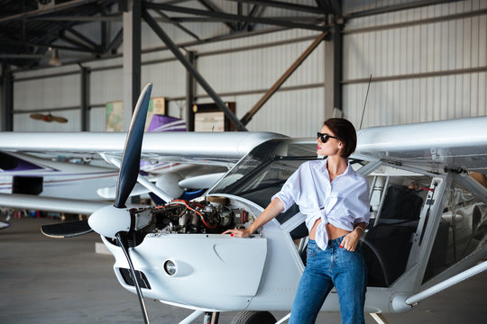 Beautiful young woman in sunglasses posing near plane in angar