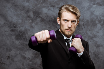 Young successful businessman posing with dumbbells over grey background.