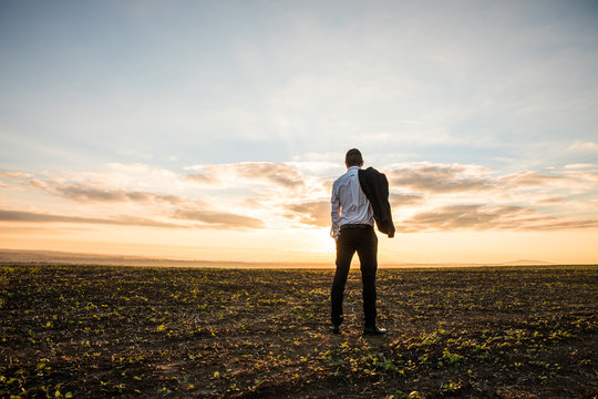 Businessman in elegant suit with his jacket hanging in field over sunset background