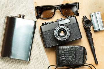 Accessories of the creative person. 35-mm film camera, exposure meter in leather case, steel flask, sunglasses and smoking set on wooden background. 