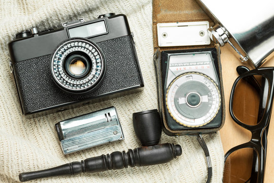 Accessories of the creative person. 35-mm film camera, exposure meter in leather case, steel flask, sunglasses and smoking set on wooden background. 