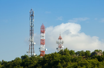 Three Telecommunication (GSM) towers with TV antennas ,satellite dishes  and repeaters at the top of a hill on a blue sky and a cloud on background