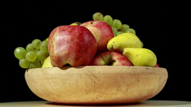 Ceramic bowl full of different fruits (apples, bananas, green grapes and pears) is rotating on black background. Loop.
