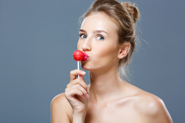 Young beautiful blonde girl kissing chupa chups over grey background.