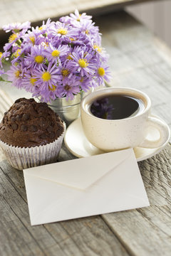 photo of sunny morning - chocolate muffin, cup of coffee and a bouquet of blue flowers and an envelope. 