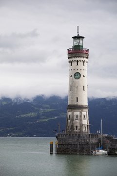 Old Lighthouse On A Pier Of A Lake With Cloud Covered Mountains In The Background; Lindau, Germany