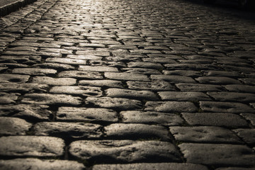Black cobbled stone road background with reflection of light seen on the road. Black or dark grey stone pavement texture.