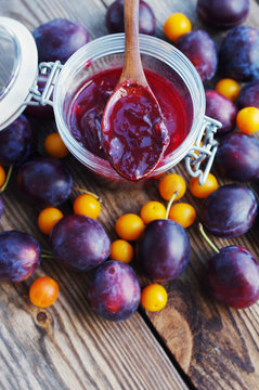 Plums and plum jam in a glass jar and a wooden spoon on a wooden surface.