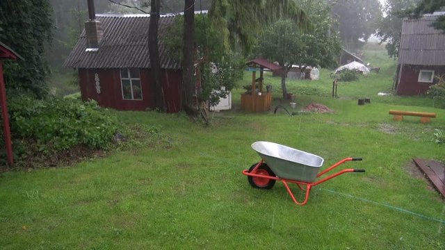 Hail and rain falling in the yard of homestead with red wooden houses and wheelbarrow