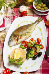 Fried gilt-head bream on a plate with a side dish of broccoli, cherry tomatoes, rice, olives and lemon.