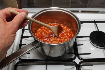 Man's hand stirring a pan of baked beans on a gas hob