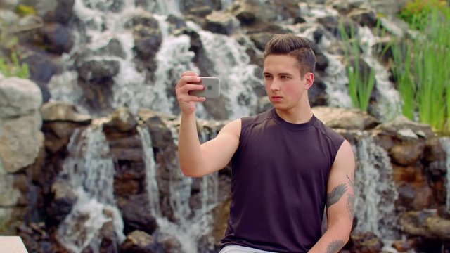 Young man taking selfie. Serious expression on man face. Man selfie. Sef photo of thoughtful man. Handsome man taking photo with phone in park. Man take photo with smartphone camera near waterfall