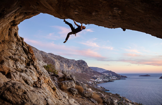 Male climber climbing along roof in cave against beautiful view of coast below 