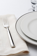 Fine dining table setting place setting with high quality white china dishes, wine glass, linen napkin and silverware fork. Selective focus on tines of fork.