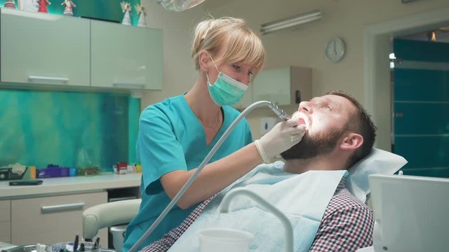Female dentist examining an drilling patient's tooth. Visit is in proffessional dental clinic. He is sitting on dental chair. He is young and has beard. Dolly and steadicam shot.
