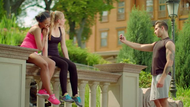 Young man taking photo of cheerful girls on phone. Group of friends taking photo on smart phone at summer. Fitness people taking photo after workout. Man taking photo of young women posing on camera