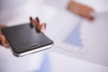 Fototapeta premium Business woman holding a smart phone and a business report in background - business concept.