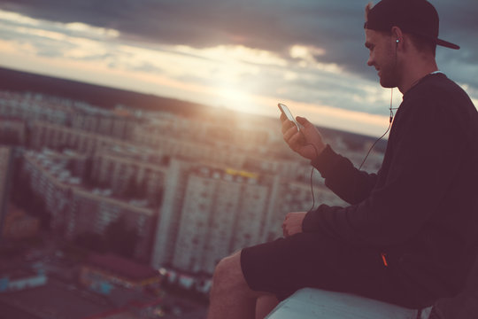 Brave smiling man sitting on the edge of the roof with smartphone and listening music