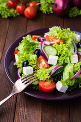 Greek salad (lettuce, tomatoes, feta cheese, cucumbers, black olives, purple onion) on dark wooden background close up. Healthy food.