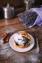 Stack of delicious pancakes with chocolate, honey, slices of banana and castor sugar on plate on wooden background, cinnamon sticks and bunch of lavender.