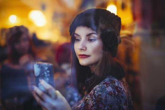 Portrait of young woman with smartphone behind windowpane of a pub in the evening