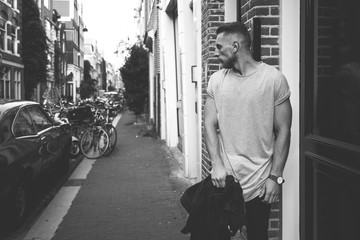 A black and white street photo of a bearded hipster guy wearing a blank gray cotton t-shirt while standing on a street background. Empty place for the logo or design. Mock up.