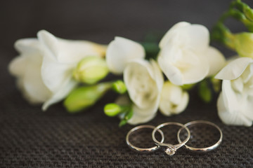 Wedding rings and engagement ring in focus, white flowers on background. Shallow focus.
