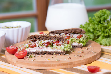 A healthy meal of whole wheat dark (brown) rye bread, curd cream cheese, radish, alfalfa sprouts, ham, cherry tomatoes and green salad on a wooden cutting board. Delicious breakfast full of vitamins.