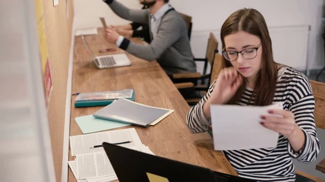 Young attractive girl is using a laptop, working with documents, making notes in the modern startup office
