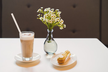 Latte in a glass and fresh eclair on dessert dish, bouquet of flowers on the background. Blank...