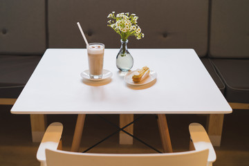 Latte in a glass and fresh eclair on dessert dish, bouquet of flowers on the background. Blank white chocolate plate on the eclair.