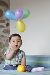 Fototapeta na wymiar baby sitting with his hand in the mouth with balloons over his head