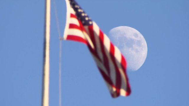 American flag waves in wind, moon in background, pull focus day time shot.