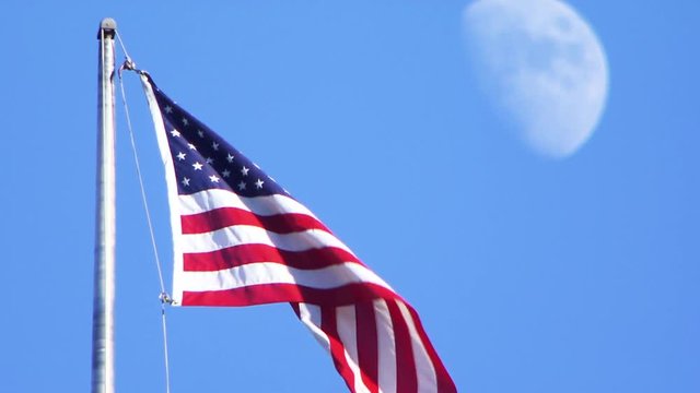 American flag waves in wind, moon in background, pull focus day time shot.