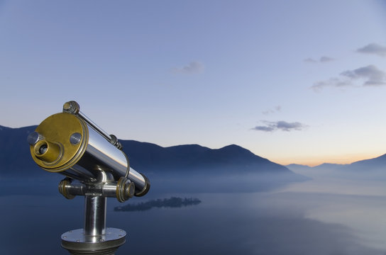 Binoculars With A View Of A Mountainous Landscape And Fog Over Lake Maggiore; Isole Di Brissago, Ticino, Switzerland